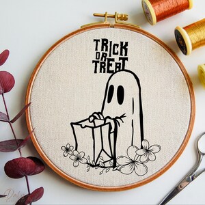 May include: A Halloween embroidery hoop with a black ghost holding a trick-or-treat bag, with floral details. The words "Trick or Treat" are in black lettering. The hoop is wooden with a beige fabric background. Threads and scissors are visible.