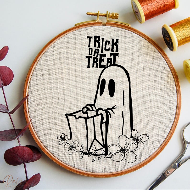 May include: A Halloween embroidery hoop with a black ghost holding a trick-or-treat bag, with floral details. The words "Trick or Treat" are in black lettering. The hoop is wooden with a beige fabric background. Threads and scissors are visible.