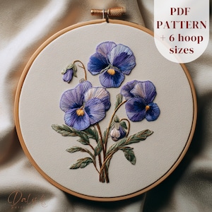 May include: An embroidery hoop featuring a detailed floral design of purple and yellow pansies. The text "PDF PATTERN + 6 hoop sizes" is visible. The embroidery is set in a wooden hoop, showcasing intricate stitching.