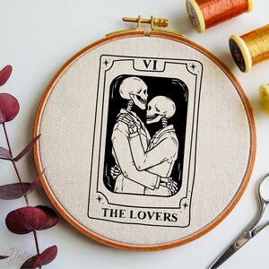May include: An embroidered hoop art piece featuring a tarot card design. The card depicts two skeletons in an embrace, with the text "VI" and "THE LOVERS". The wooden hoop holds beige fabric, with spools of thread visible.