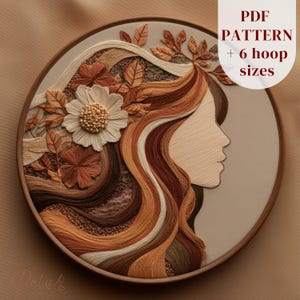 May include: A round embroidery hoop featuring a profile of a woman with flowing hair in shades of brown, beige, and orange. The design includes a large white flower and leaves. The text "PDF PATTERN + 6 hoop sizes" is visible.