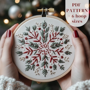 May include: Embroidered snowflake design in a wooden hoop. The snowflake features red, green, and cream-colored thread on a white background. The text "PDF PATTERN + 6 hoop sizes" is in the upper right corner. The design is held by hands with red nail polish.