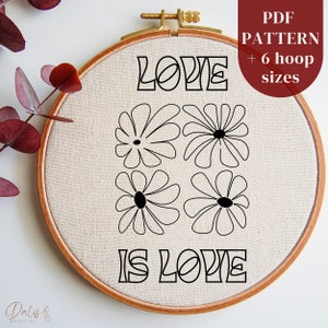 Könnte beinhalten: Ein schwarz-weißes Stickmuster mit dem Text "LOVE IS LOVE" in stilisierter Schrift und vier einfachen Blumenmotiven. Das Muster ist in einem hölzernen Stickrahmen dargestellt.
