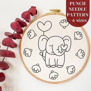 Peut inclure: Un motif de broderie au point de croix sur toile ronde, représentant un éléphant de dessin animé tenant un ballon en forme de cœur. Le motif est entouré de formes florales et encadré dans un cercle à broder en bois. Texte : "PUNCH NEEDLE PATTERN + 6 sizes."