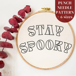 Puede incluir: Un aro de bordado blanco con el texto "STAY SPOOKY" en letras negras delineadas. El aro está sobre un fondo blanco con una rama de hojas rojas.