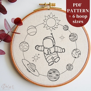 Op de afbeelding: Een zwart-wit borduurpatroon van een astronaut die in de ruimte zweeft met planeten en sterren. Het patroon is op een beige stof in een houten borduurring. De tekst "PDF PATTERN + 6 hoop sizes" staat in de rechterbovenhoek van de afbeelding.