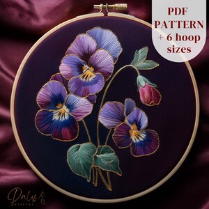 May include: A detailed embroidery of pansies in shades of purple, blue, and yellow, set against a dark purple background. The floral design is framed within a wooden embroidery hoop. Text on the image reads "PDF PATTERN + 6 hoop sizes."