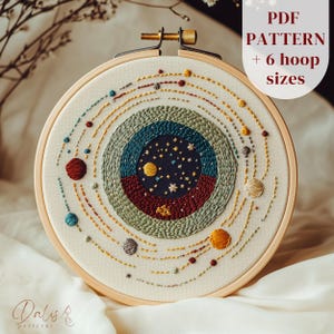 Könnte beinhalten: Gesticktes Stickrahmen-Kunstwerk mit einem Himmelsdesign mit Planeten und Sternen. Das Kunstwerk befindet sich in einem Holzreifen und enthält den Text "PDF PATTERN + 6 hoop sizes". Die Farbpalette umfasst Blau-, Rot-, Gelb- und Grüntöne.