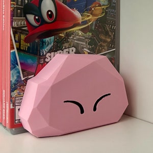 May include: A pink, cloud-shaped, 3D printed object with a cartoon face. The object is sitting on a white shelf next to a stack of video game cases featuring Mario and a blue Nintendo Switch controller.