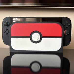 以下が含まれることがあります： 赤、白、黒のポケモンをテーマにしたケースが付いた黒色のNintendo Switchコンソール。ケースはモンスターボールに似せてデザインされています。コンソールは反射面に置かれています。