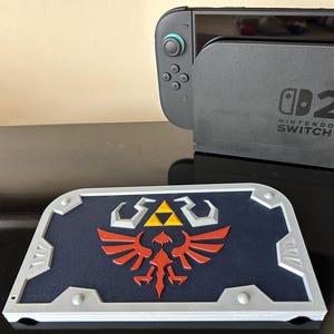 May include: A blue, red, yellow, and silver shield featuring the Hylian Crest from The Legend of Zelda. The shield is placed in front of a Nintendo Switch console and dock.