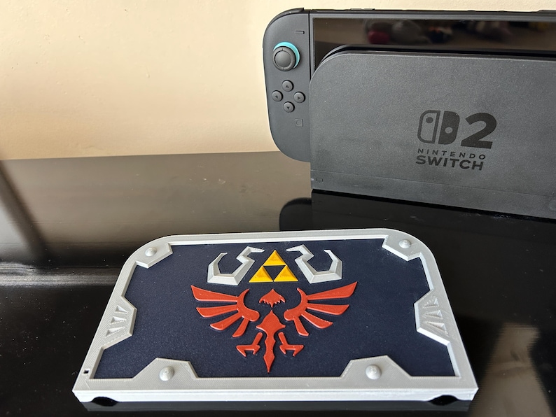 May include: A blue, red, yellow, and silver shield featuring the Hylian Crest from The Legend of Zelda. The shield is placed in front of a Nintendo Switch console and dock.