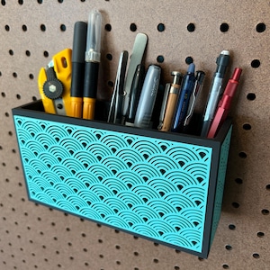May include: A teal and black desk organizer with a wave pattern, holding various pens, pencils, and tools. The organizer is mounted on a brown pegboard, providing a stylish storage solution for office or craft supplies.