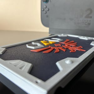 May include: A shield-shaped case in grey and white, featuring a red, yellow, and white emblem, designed for a Nintendo Switch console. The case sits on a reflective black surface, with the console visible in the background.