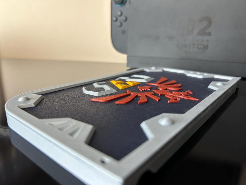 May include: A shield-shaped case in grey and white, featuring a red, yellow, and white emblem, designed for a Nintendo Switch console. The case sits on a reflective black surface, with the console visible in the background.