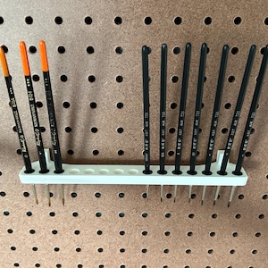 May include: White plastic paintbrush holder with 12 slots, mounted on a pegboard wall. The holder is filled with black and orange paintbrushes.