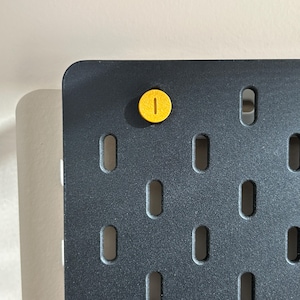 May include: A dark gray pegboard with evenly spaced, oblong holes. A yellow peg is inserted into one of the holes. The pegboard has a rounded corner and is set against a light-colored background. The peg is a bright yellow color.