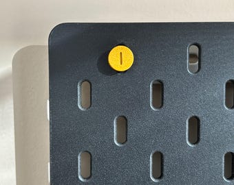 May include: A dark gray pegboard with evenly spaced, oblong holes. A yellow peg is inserted into one of the holes. The pegboard has a rounded corner and is set against a light-colored background. The peg is a bright yellow color.