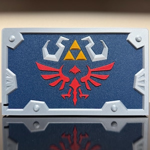 May include: A rectangular, blue and silver decorative item featuring a red emblem and a gold triangle. The item has a silver border with silver corner accents. The emblem is a stylized bird with outstretched wings.