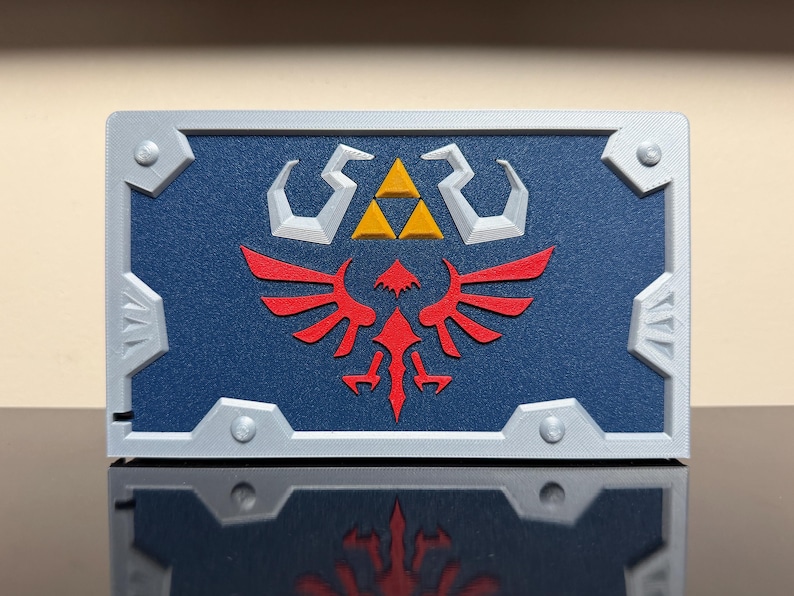 May include: A rectangular, blue and silver decorative item featuring a red emblem and a gold triangle. The item has a silver border with silver corner accents. The emblem is a stylized bird with outstretched wings.