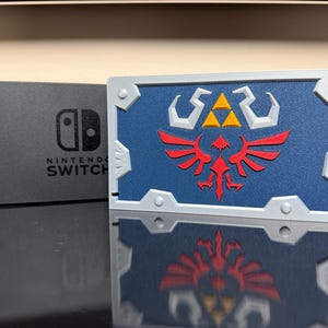 May include: A Nintendo Switch console and a blue and silver decorative shield. The shield features a red and yellow emblem on a blue background. The console is black with the words "Nintendo Switch" printed on it.