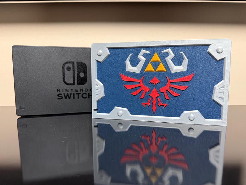May include: A Nintendo Switch console and a blue and silver decorative shield. The shield features a red and yellow emblem on a blue background. The console is black with the words "Nintendo Switch" printed on it.