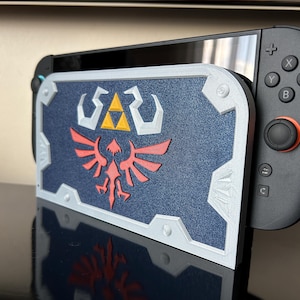 May include: A navy blue and white 3D-printed shield-shaped stand for a Nintendo Switch. The shield features a red and white emblem with a yellow triangle. The Switch's black controller is visible on the right.