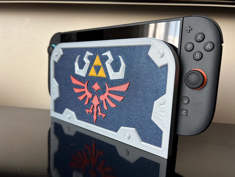 May include: A navy blue and white 3D-printed shield-shaped stand for a Nintendo Switch. The shield features a red and white emblem with a yellow triangle. The Switch's black controller is visible on the right.