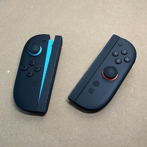 May include: Two black game controllers with buttons and joysticks. One controller has a blue accent, the other has an orange accent. The controllers are designed for gaming.