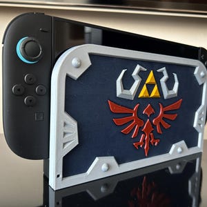 May include: A black Nintendo Switch console with a custom shield-shaped case. The case is navy blue with a silver border and features a red and gold emblem. The console is on a reflective surface.