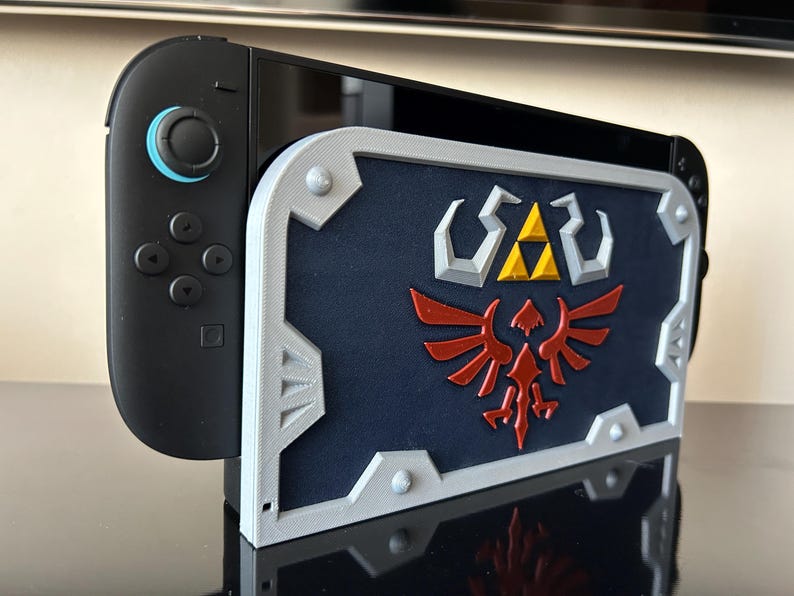 May include: A black Nintendo Switch console with a custom shield-shaped case. The case is navy blue with a silver border and features a red and gold emblem. The console is on a reflective surface.