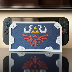 May include: A blue and white shield-shaped case with a red and yellow emblem, designed for a Nintendo Switch console. The case has a textured surface and is positioned in front of the console, with the controllers visible on either side.