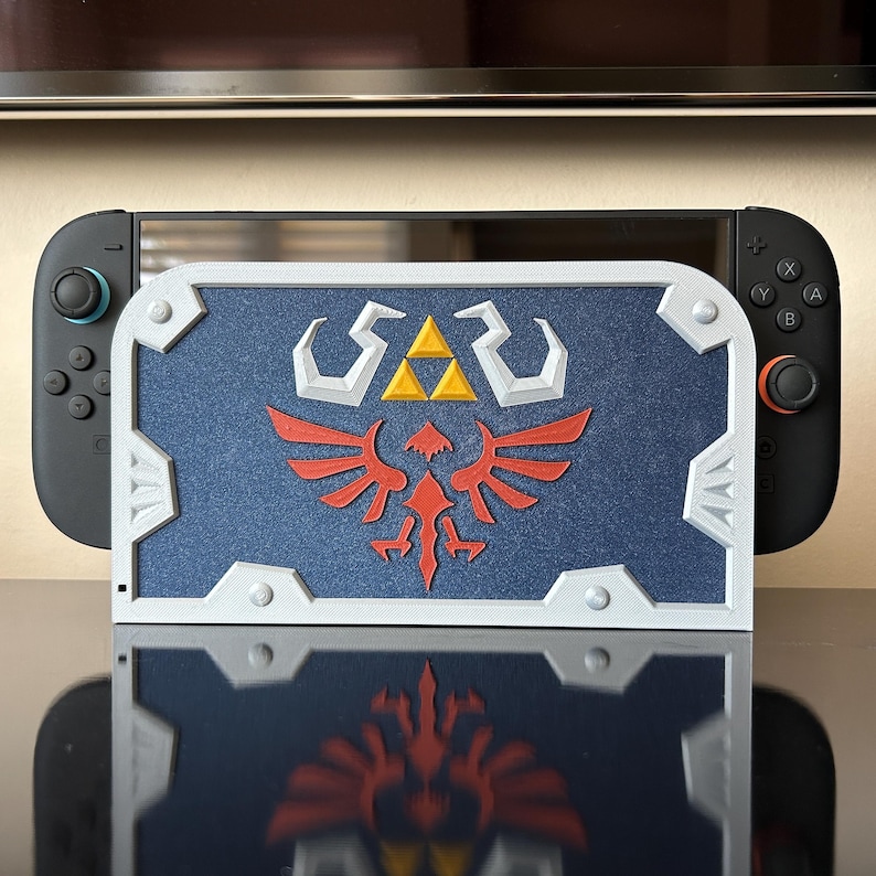 May include: A blue and white shield-shaped case for a Nintendo Switch. The case features a red and yellow emblem with the text "SAR". The Nintendo Switch is black with teal and black controllers.