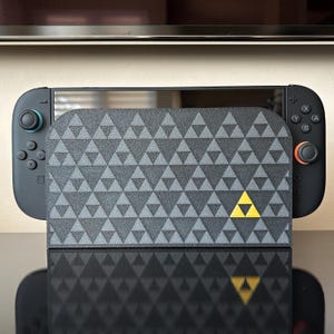 May include: A black Nintendo Switch console with a patterned cover featuring a repeating triangle design in shades of gray. The cover has a yellow triangle emblem. The console has teal and orange accents.