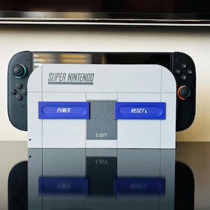 May include: A grey Super Nintendo Switch dock featuring blue and grey buttons labelled "POWER" and "RESET". The dock is in front of a black Nintendo Switch console with blue and orange accents.