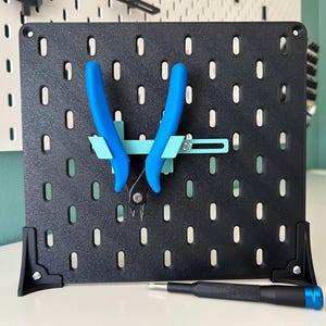 May include: A black pegboard with a blue and turquoise tool holder and a blue screwdriver on a white surface.