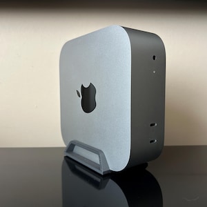 May include: A silver and black Apple Mac Mini computer with the Apple logo on the front. The computer is standing upright in a grey stand. The computer has a rounded rectangular shape.