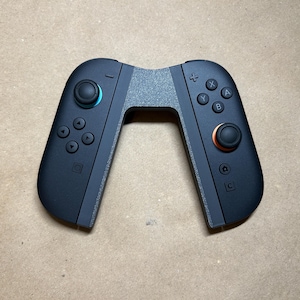 以下が含まれることがあります： 黒いゲームコントローラーと灰色の接続部分。 コントローラーには、ボタン、方向パッド、青とオレンジのアクセントが付いたジョイスティックがあります。 ゲーム用に設計されています。