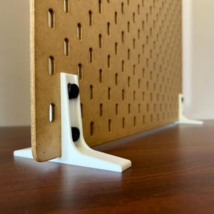 May include: A brown pegboard with evenly spaced slots, supported by white plastic feet. The pegboard is designed to hold various items. The white feet have black rubber bumpers. The pegboard is on a dark brown surface.