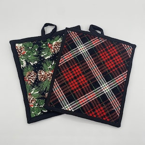 May include: Two square pot holders with black trim and hanging loops. One features a black background with pine needles, pine cones, and red berries. The other has a red, black, and white plaid pattern.