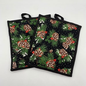 May include: Two square quilted potholders featuring a black background with a repeating pattern of brown pinecones, green pine needles, and small red berries. Each potholder has a solid black fabric border and a small black fabric hanging loop.