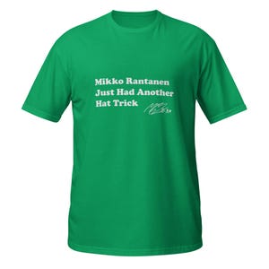 Op de afbeelding: Een groen t-shirt met witte tekst die luidt: "Mikko Rantanen Just Had Another Hat Trick" en een handtekening. Het shirt is effen, heldergroen van kleur.