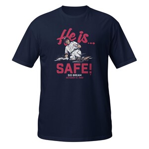 Puede incluir: Una camiseta azul marino con un gráfico rojo de un jugador de béisbol deslizándose hacia el plato de home. El gráfico dice "He is... SAFE!" con el nombre del jugador "Sid Bream" y la fecha "October 14, 1992" debajo.