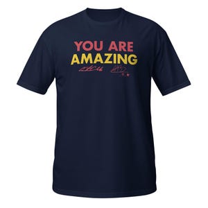May include: Navy blue t-shirt with the text "YOU ARE AMAZING" in yellow and red lettering. The shirt features a red signature.