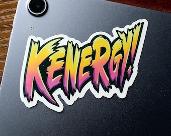 May include: A sticker with the word "Kenergy!" in a stylized font. The letters are yellow, orange, and pink with black outlines. The sticker is on a gray background.