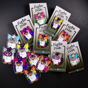 Può includere: Una collezione di spille smaltate colorate con il giocattolo Furby con il testo "Furbies with Pride" su ogni carta. Le spille sono arcobaleno e presentano diverse espressioni sui volti dei Furby.