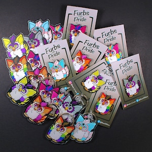 Op de afbeelding: Een verzameling kleurrijke Furby-sleutelhangers met regenboogkleuren van de pride-vlag. De sleutelhangers tonen verschillende Furby-ontwerpen met verschillende uitdrukkingen. De sleutelhangers worden getoond op een zwarte achtergrond met kaarten waarop "Furbies with Pride" staat.