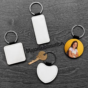 May include: A collection of keychains with silver rings and black leather accents. The keychains have various shapes: a rectangle, a square, a heart, and a circle with a photo of a girl. The keychains are blank and ready for customization.