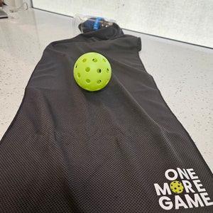 May include: A black microfiber towel with the words "ONE MORE GAME" and a small yellow pickleball printed on it. A yellow pickleball is sitting on top of the towel.