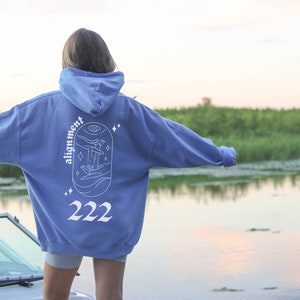 May include: A light blue hoodie with a white graphic design on the back. The design features a hand reaching towards a celestial eye with stars and waves. The text "alignment" and "222" are also included in the design.